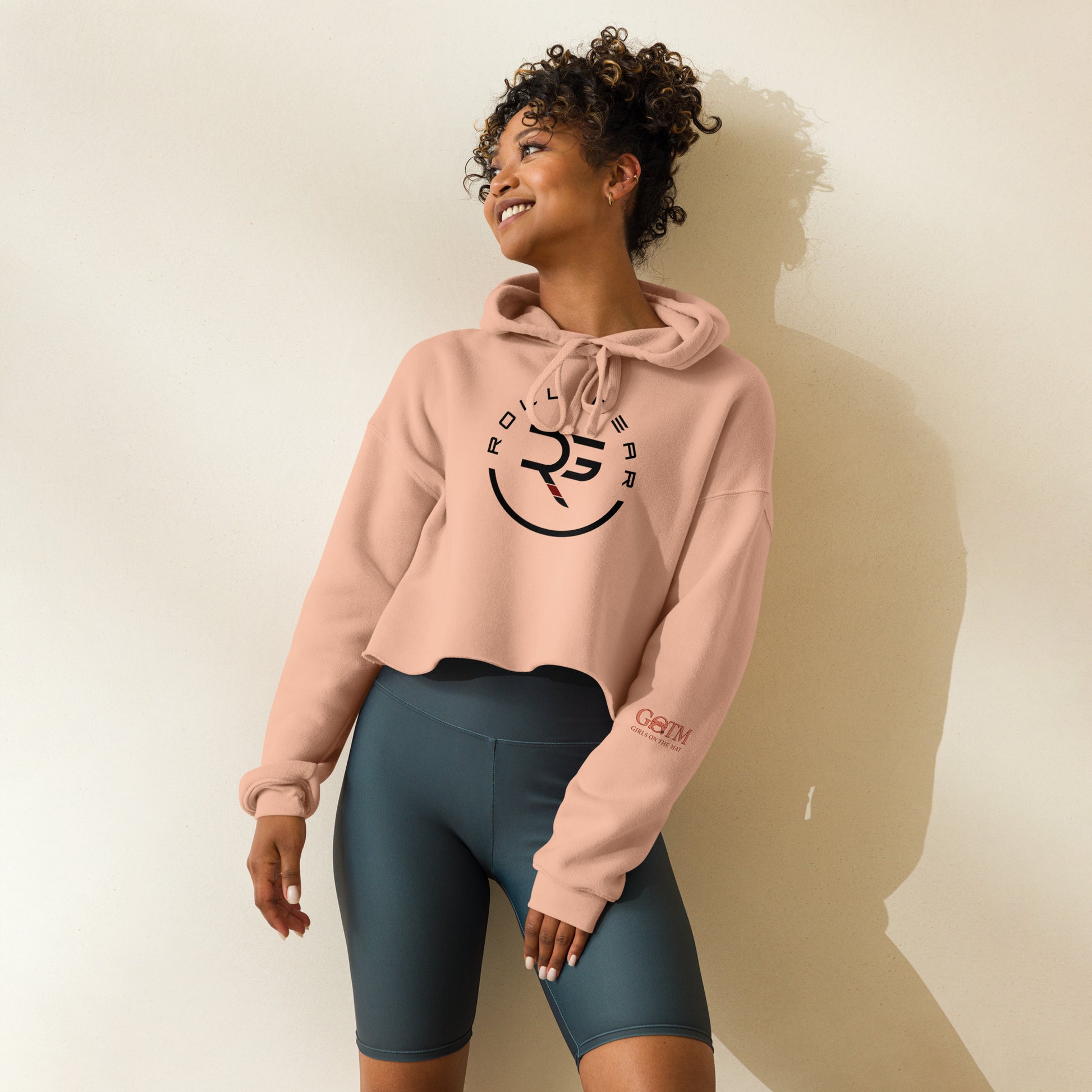 Woman wearing a pink hoodie with a logo and dark shorts on a plain background