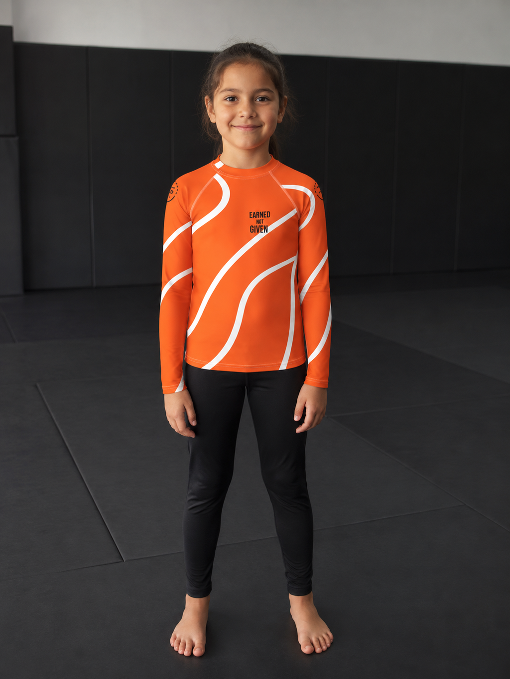 Child wearing an orange and white long-sleeve rash guard with black pants on a dark background