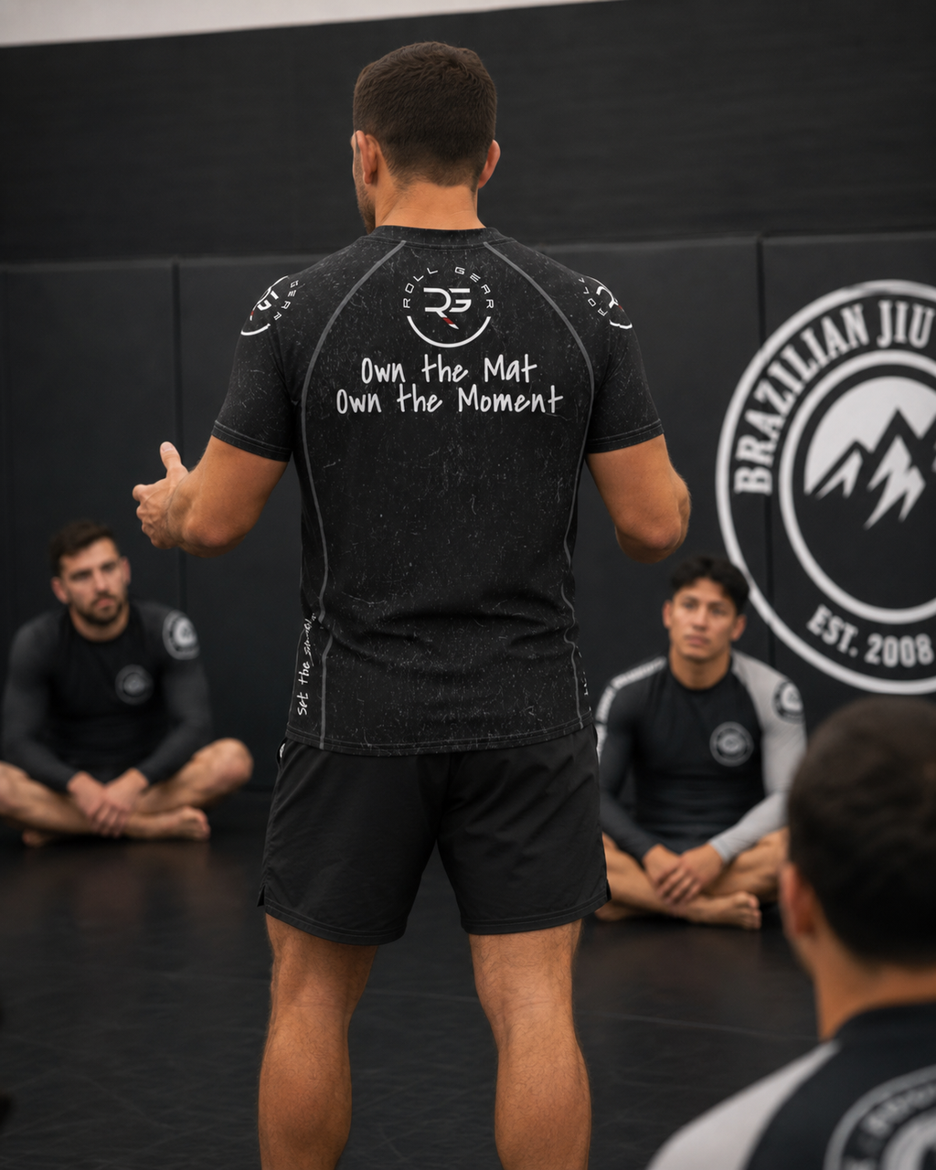 Person in a black athletic outfit with text on the back, standing in front of a group in a Brazilian Jiu-Jitsu setting.