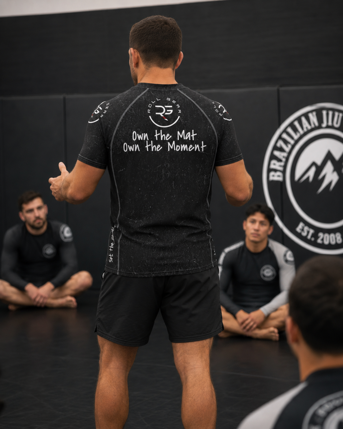 Person in a black athletic outfit with text on the back, standing in front of a group in a Brazilian Jiu-Jitsu setting.