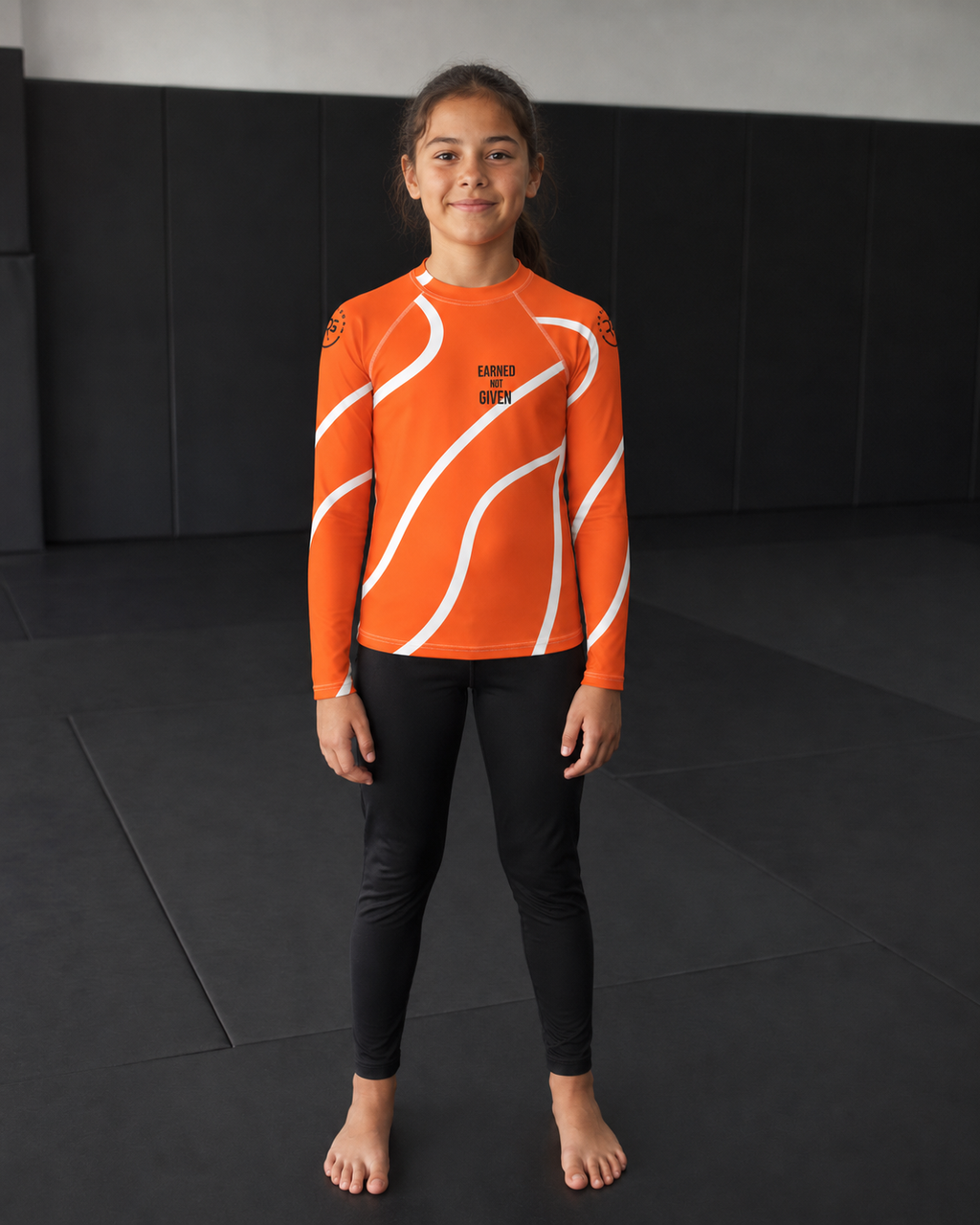 Child wearing an orange and white long-sleeve rash guard and black pants in a martial arts dojo.