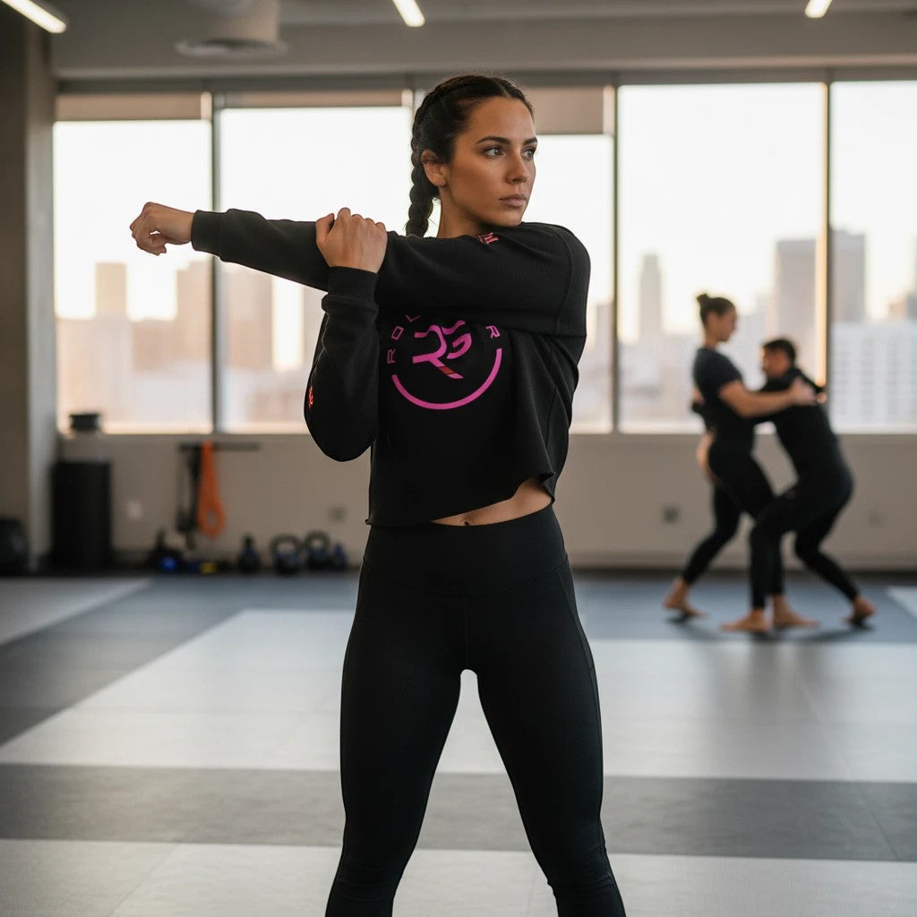 Roll Gear black cropped hoodie with pink logo, worn by a woman stretching in a gym. Women's BJJ apparel.