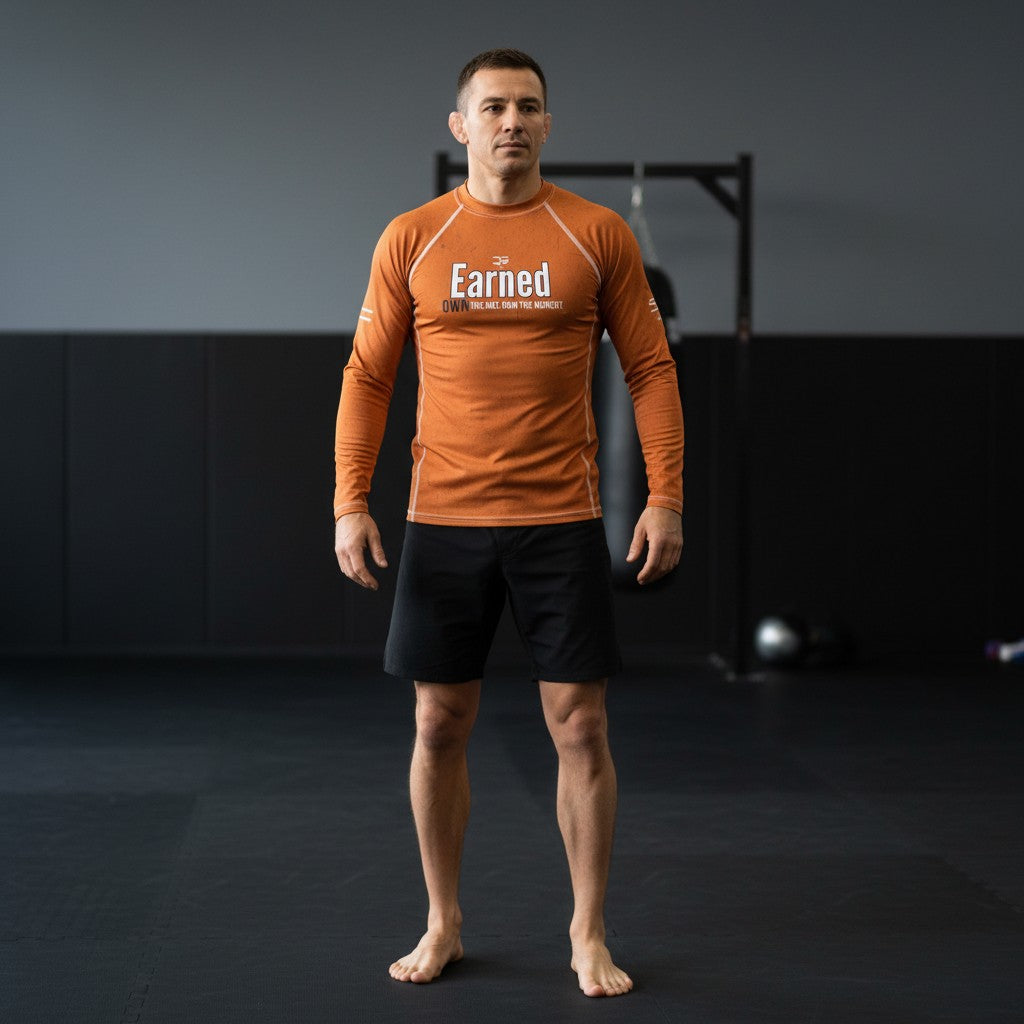 Roll Gear youth long sleeve rash guard in burnt orange with "Earned Not Given" graphic. Moisture-wicking, 4-way stretch fabric for BJJ, wrestling, and MMA.